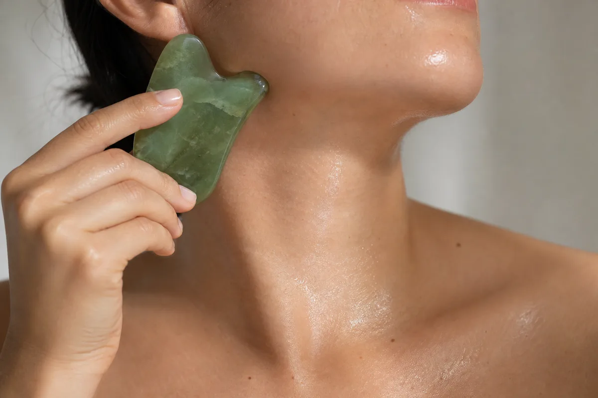 Main faisant glisser une pierre de gua sha en jade le long de la mâchoire et du cou, sans visage visible