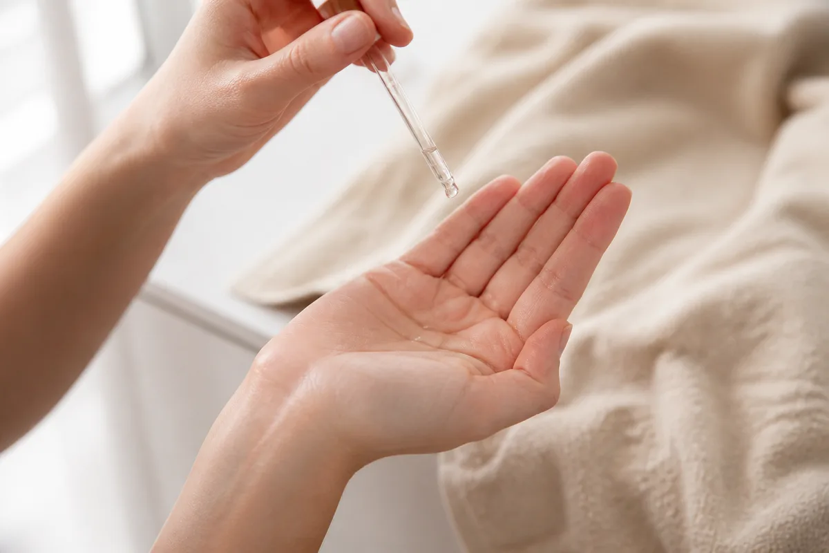 Main appliquant une goutte de sérum au bout des doigts au-dessus d’une pipette en verre, serviette de lin beige